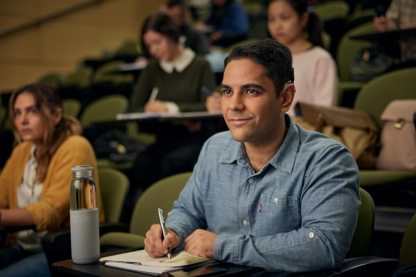 A man sitting listening to a lecture and taking notes while displaying his cochlear implant